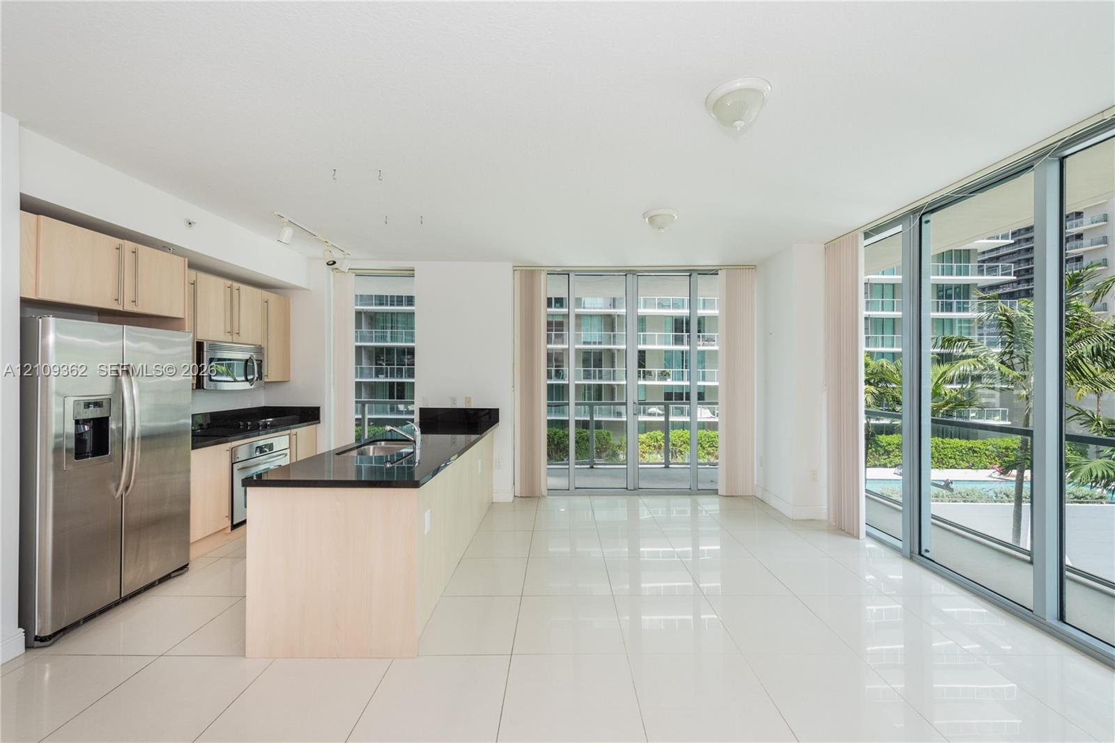 Kitchen area featuring light-finish cabinetry, dark solid-surface countertops, stainless steel appliances, floor-to-ceiling glass sliding doors, and light-toned tile flooring