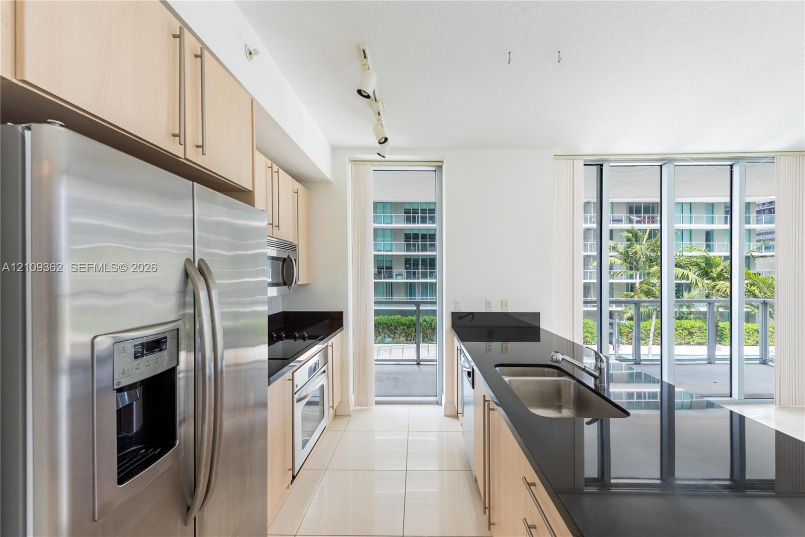 Kitchen featuring light wood-finish cabinetry, black stone countertops, stainless steel appliances, tile flooring, and floor-to-ceiling windows