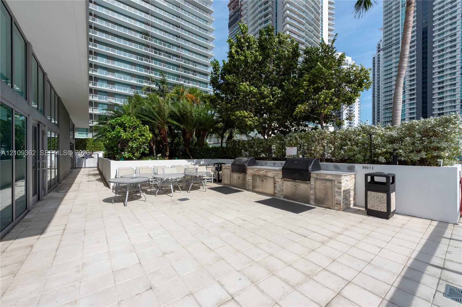 Outdoor terrace featuring light-toned tile flooring, built-in outdoor kitchen with stone veneer base, integrated stainless steel grills, and mature landscaping