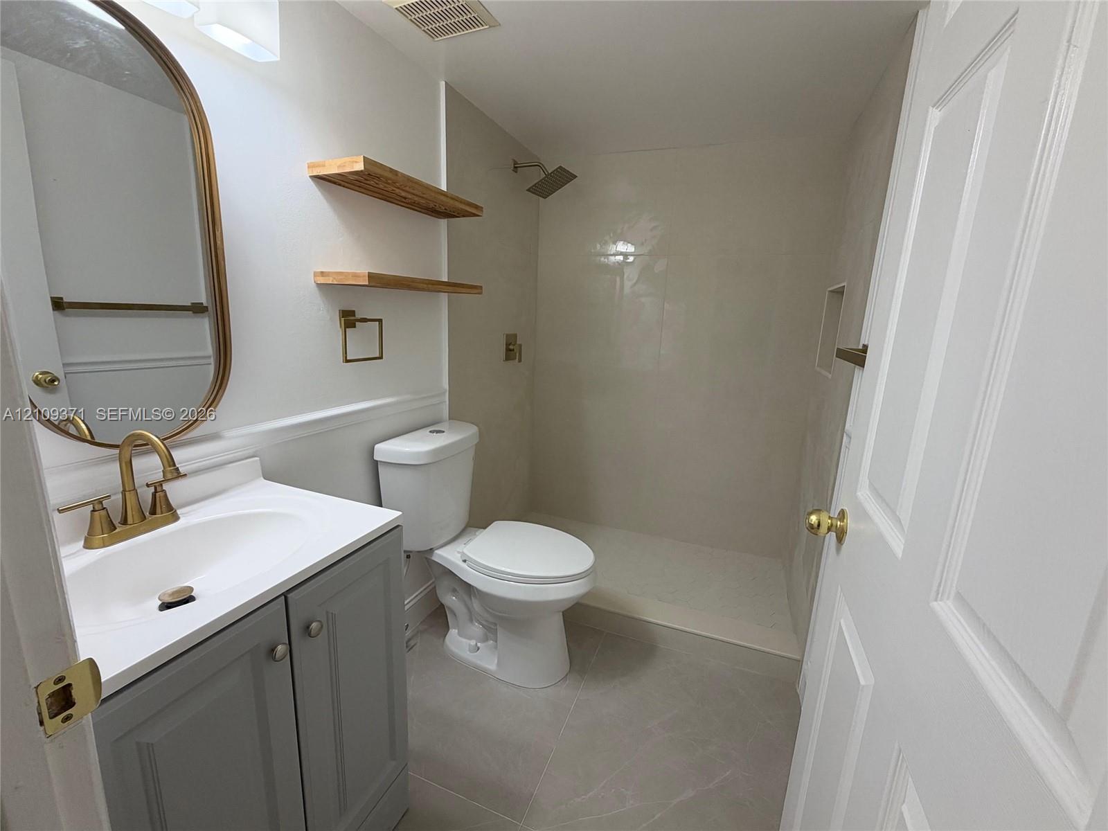 Bathroom featuring a grey vanity with a white countertop, brass-finish faucet and hardware, walk-in shower with large-format tile surround, and floating wood shelving
