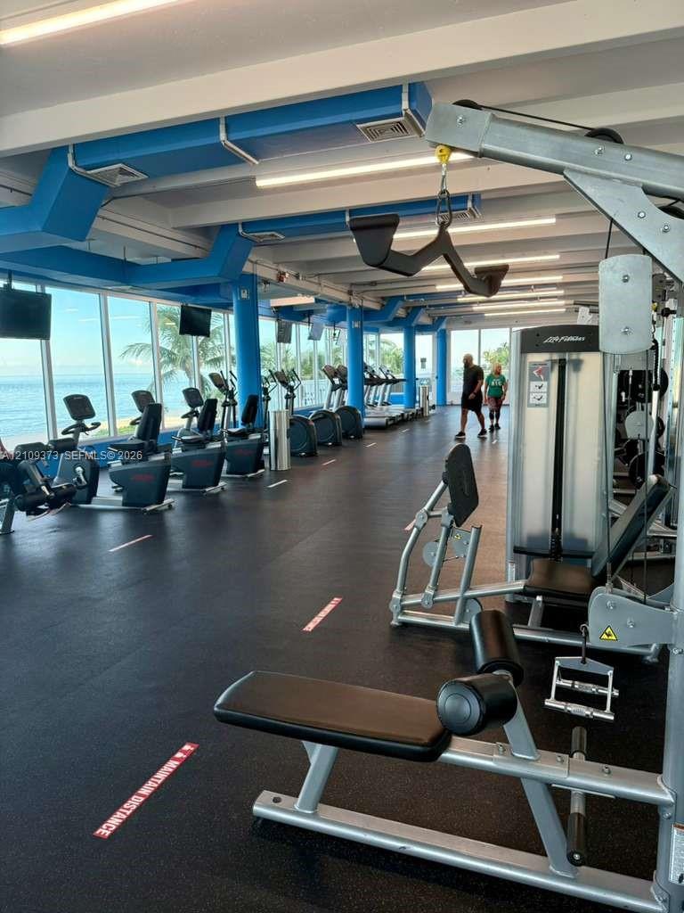 Fitness center featuring rubber flooring, exposed industrial-style blue ceiling ductwork, and floor-to-ceiling windows with coastal scenery