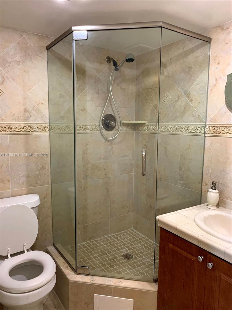 Corner glass-enclosed shower with a handheld fixture, decorative tile backsplash, integrated shelving, and neutral-tone stone-finish wall tile
