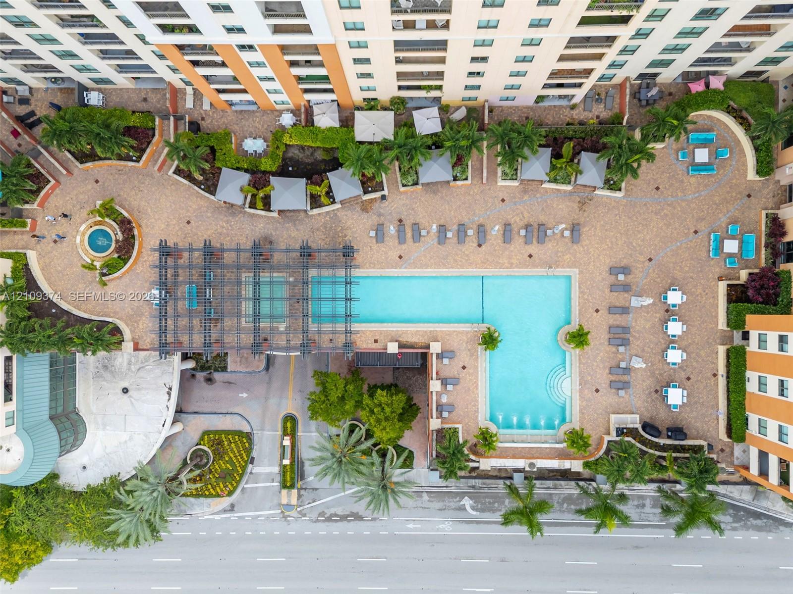 Aerial perspective of a multi-level complex featuring an L-shaped swimming pool, paved sundeck, decorative pergola, manicured landscaping, and a circular fountain