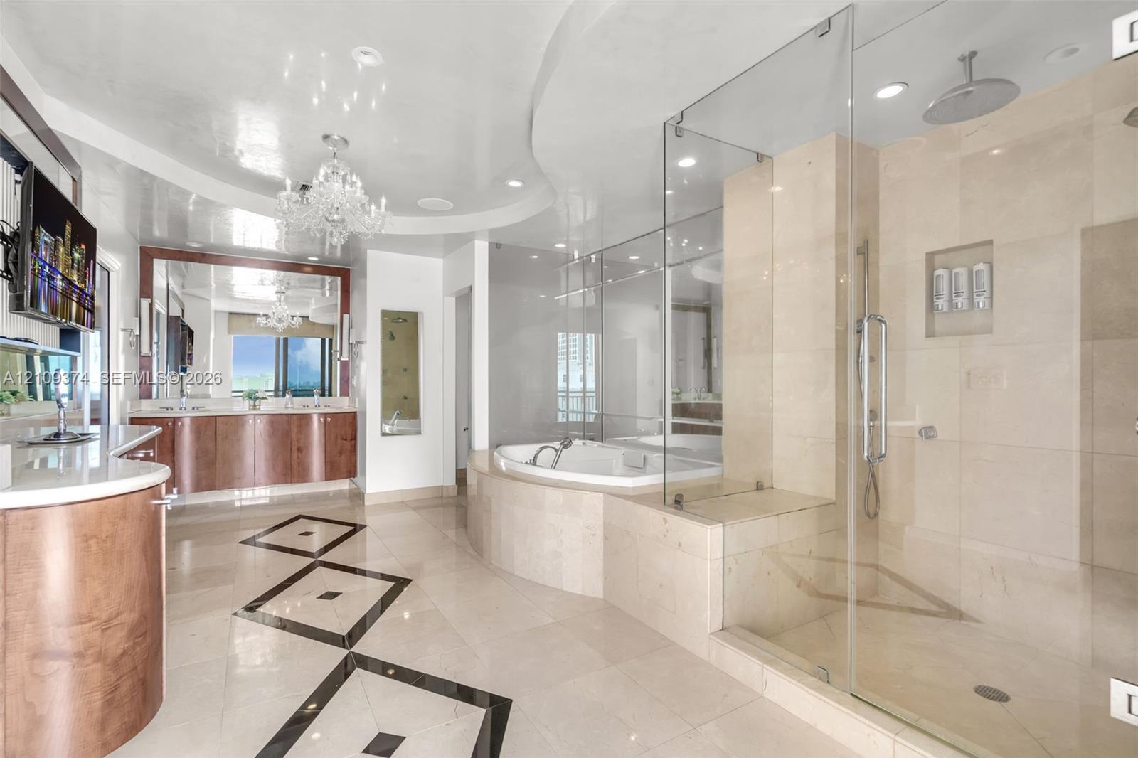 Bathroom featuring stone tile flooring with decorative inlay, a glass-enclosed shower with integrated shelving, a built-in soaking tub, wood-finish cabinetry with stone surfaces, and a ceiling-mounted crystal chandelier