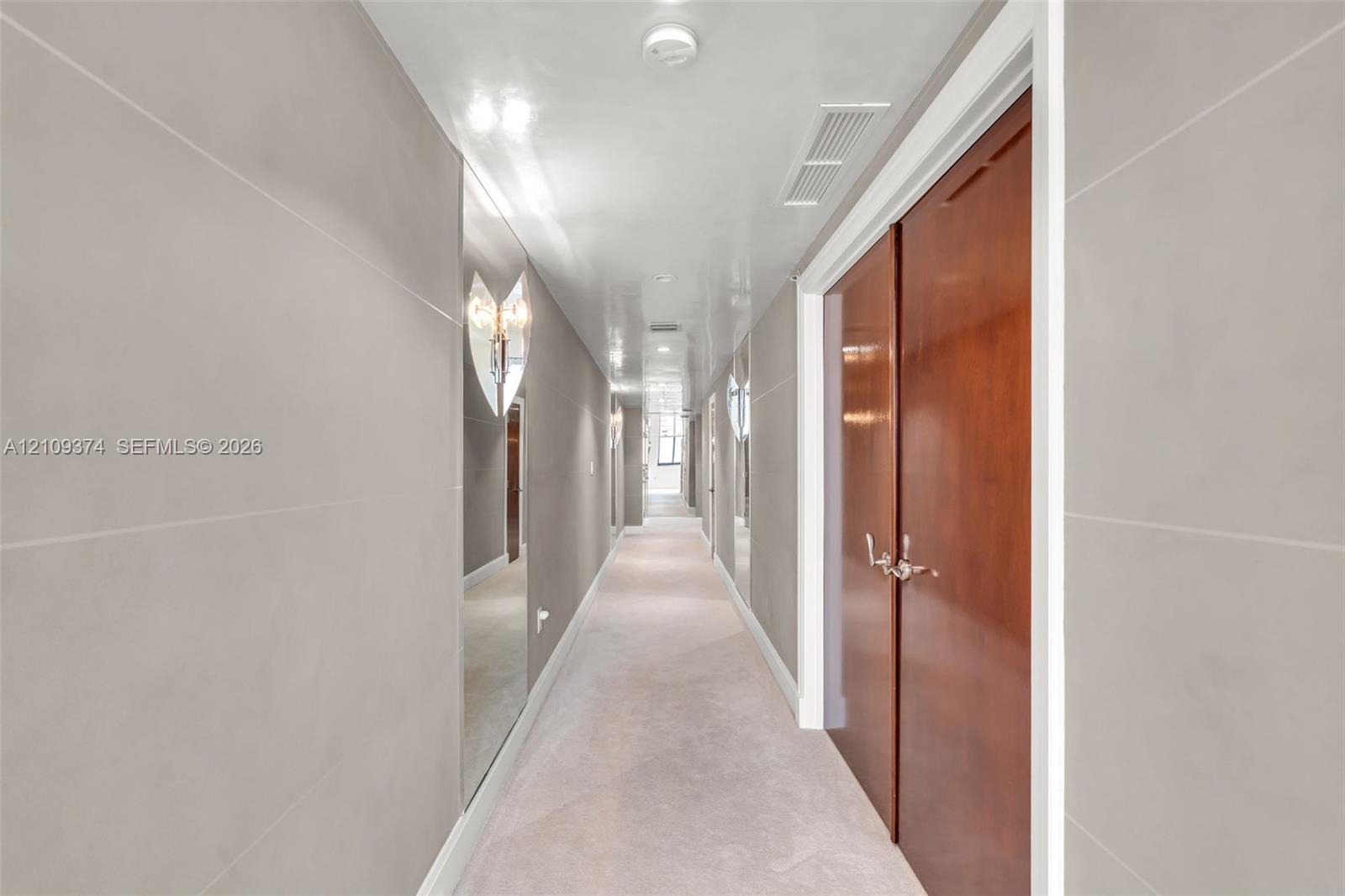 Hallway featuring large-format light gray wall panels, full-height wall mirrors, double-door wood cabinetry, and recessed lighting