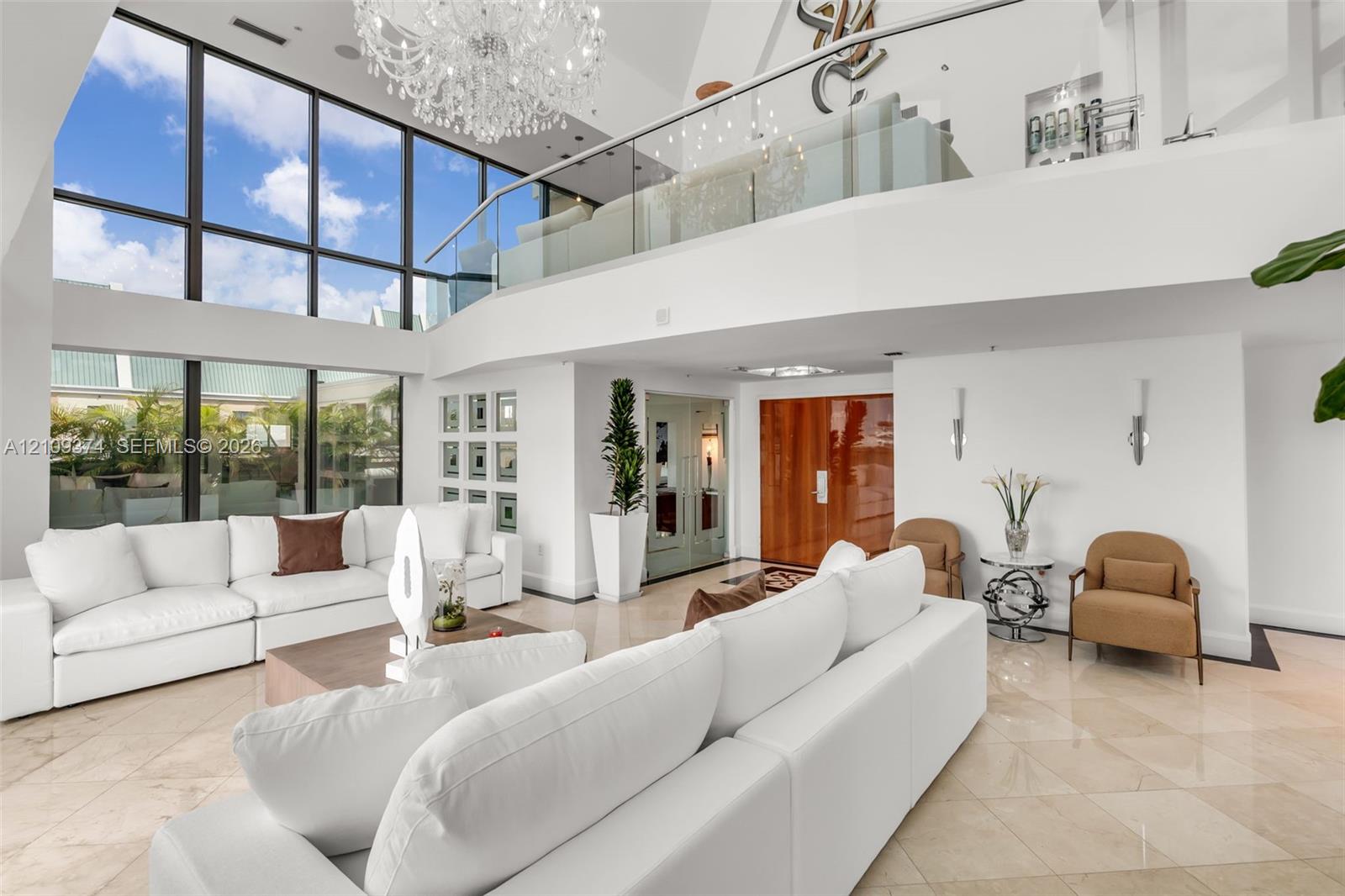 Double-height living area featuring floor-to-ceiling windows, polished stone flooring, a glass mezzanine railing, and a grand crystal chandelier Double-height living area featuring floor-to-ceiling windows, polished stone flooring, a glass mezzanine railing, and a grand crystal chandelier