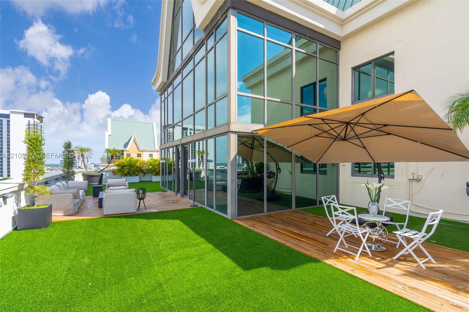 Expansive rooftop terrace featuring floor-to-ceiling glass wall panels, synthetic turf lawn, wood-finish decking, and light-colored stucco exterior siding