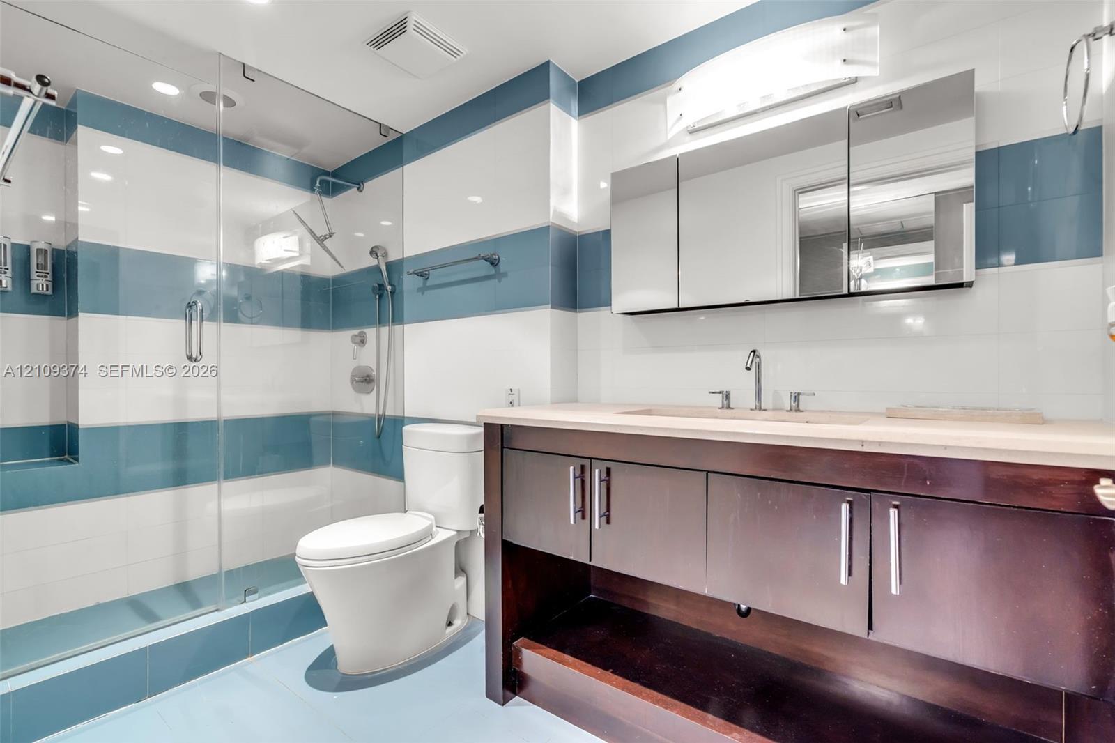 Bathroom featuring a glass-enclosed shower, wood-finish vanity with stone countertop, integrated undermount sink, and multi-tone horizontal wall tiling