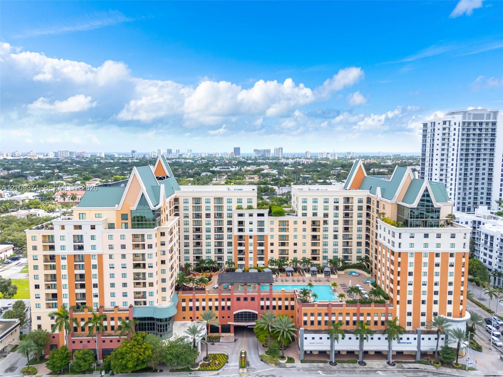 Multi-story high-rise building featuring a structured facade, gabled rooflines, recessed balconies, an elevated outdoor swimming pool, and manicured tropical landscaping