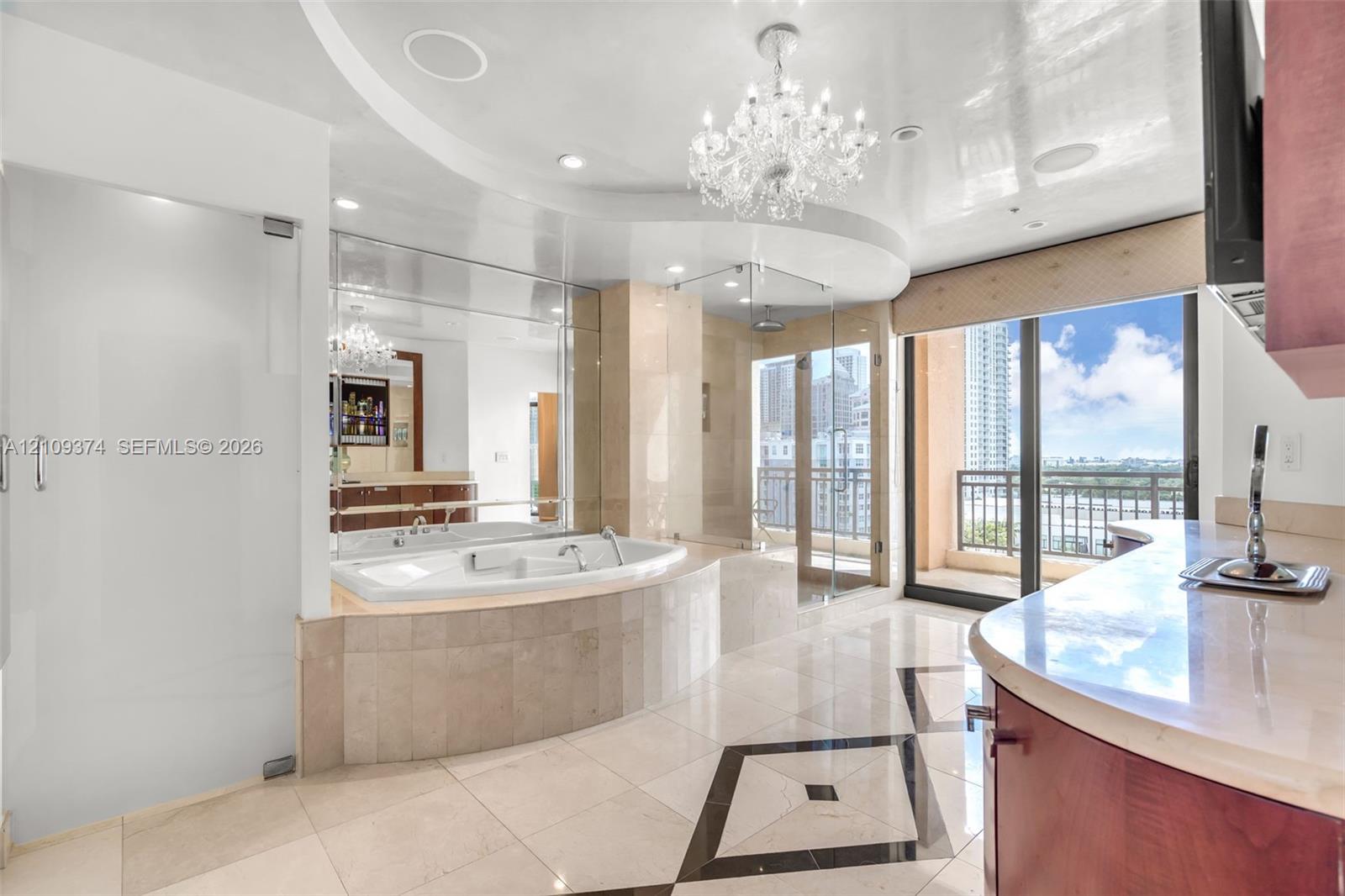 Soaking tub with stone surround, glass-enclosed shower, expansive vanity with stone countertop, ornate chandelier, and polished tile flooring with geometric inlay