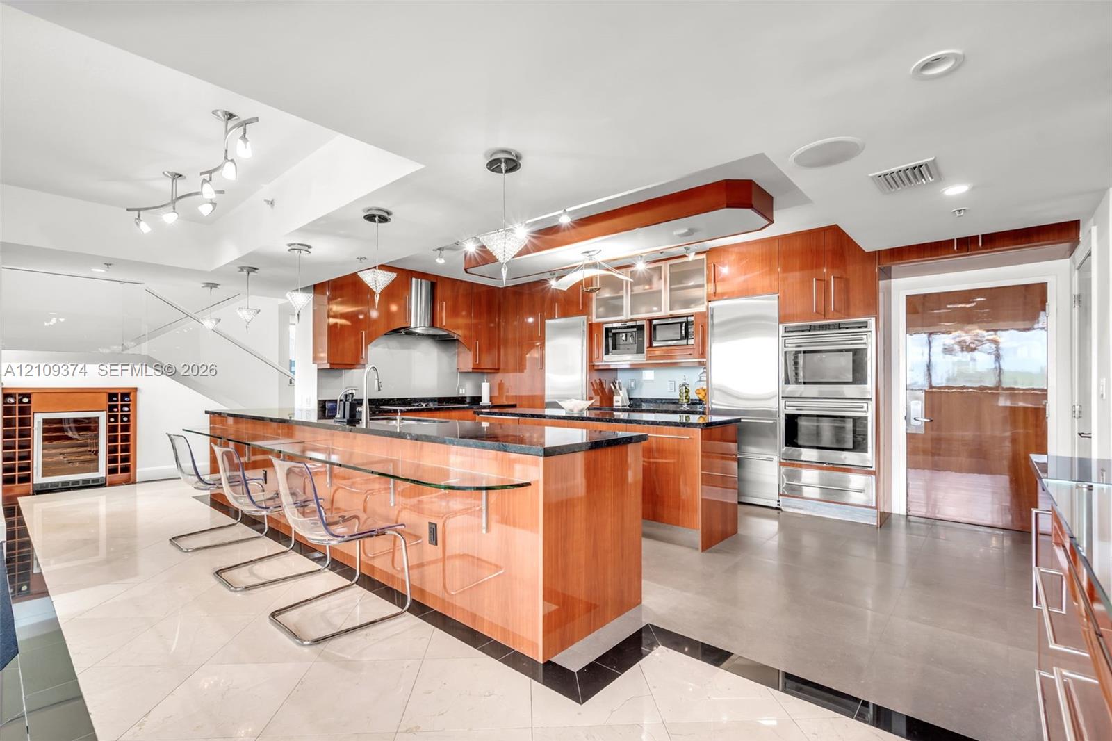 Gourmet kitchen featuring high-gloss wood cabinetry, dark stone countertops, integrated stainless steel appliances, a glass-top breakfast bar, and polished tile flooring