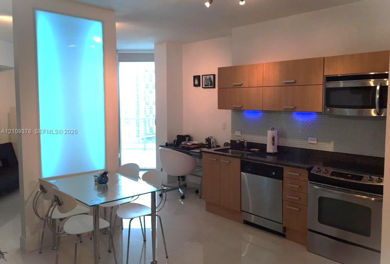 Kitchen featuring wood-finish cabinetry, stainless steel appliances, dark stone-surface countertops, glass tile backsplash with integrated under-cabinet lighting, and an illuminated wall panel
