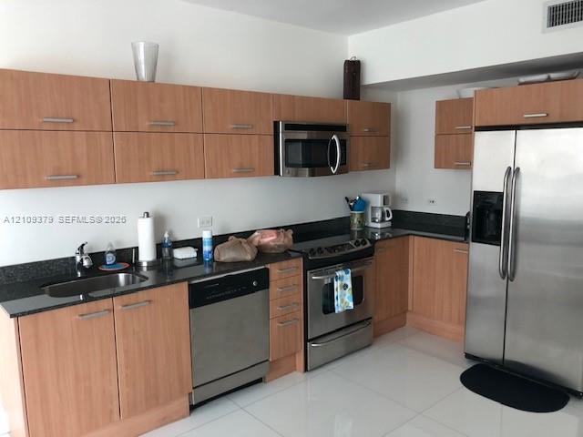 Kitchen featuring wood-finish cabinetry, dark stone countertops, stainless steel appliances, and large-format tile flooring