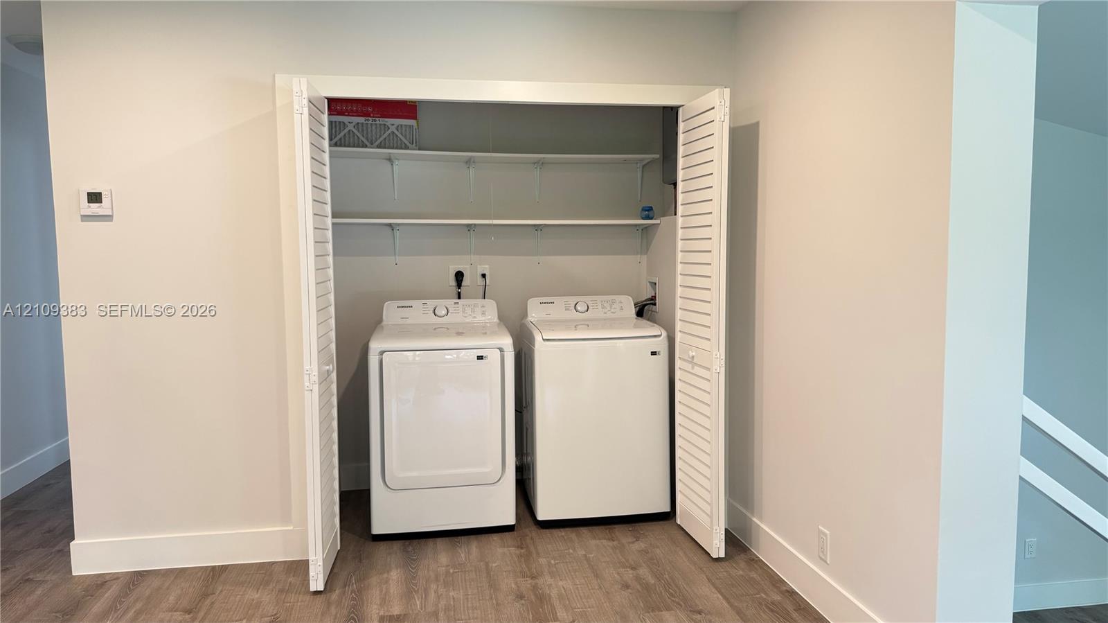 Laundry closet featuring white louvered bifold doors, wood-finish flooring, and built-in upper shelving