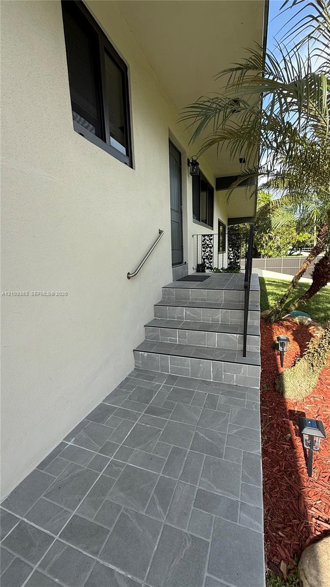 Exterior entryway featuring stone-tiled steps, a brushed metal handrail, a smooth stucco facade, and dark-framed windows