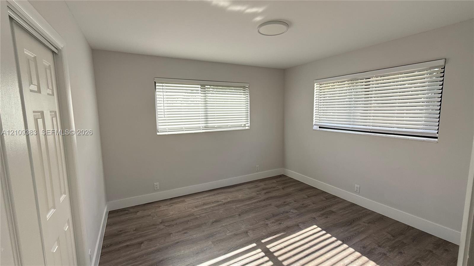 Wood-finish flooring, neutral wall tones, two windows with horizontal blinds, and a flush-mount overhead light fixture