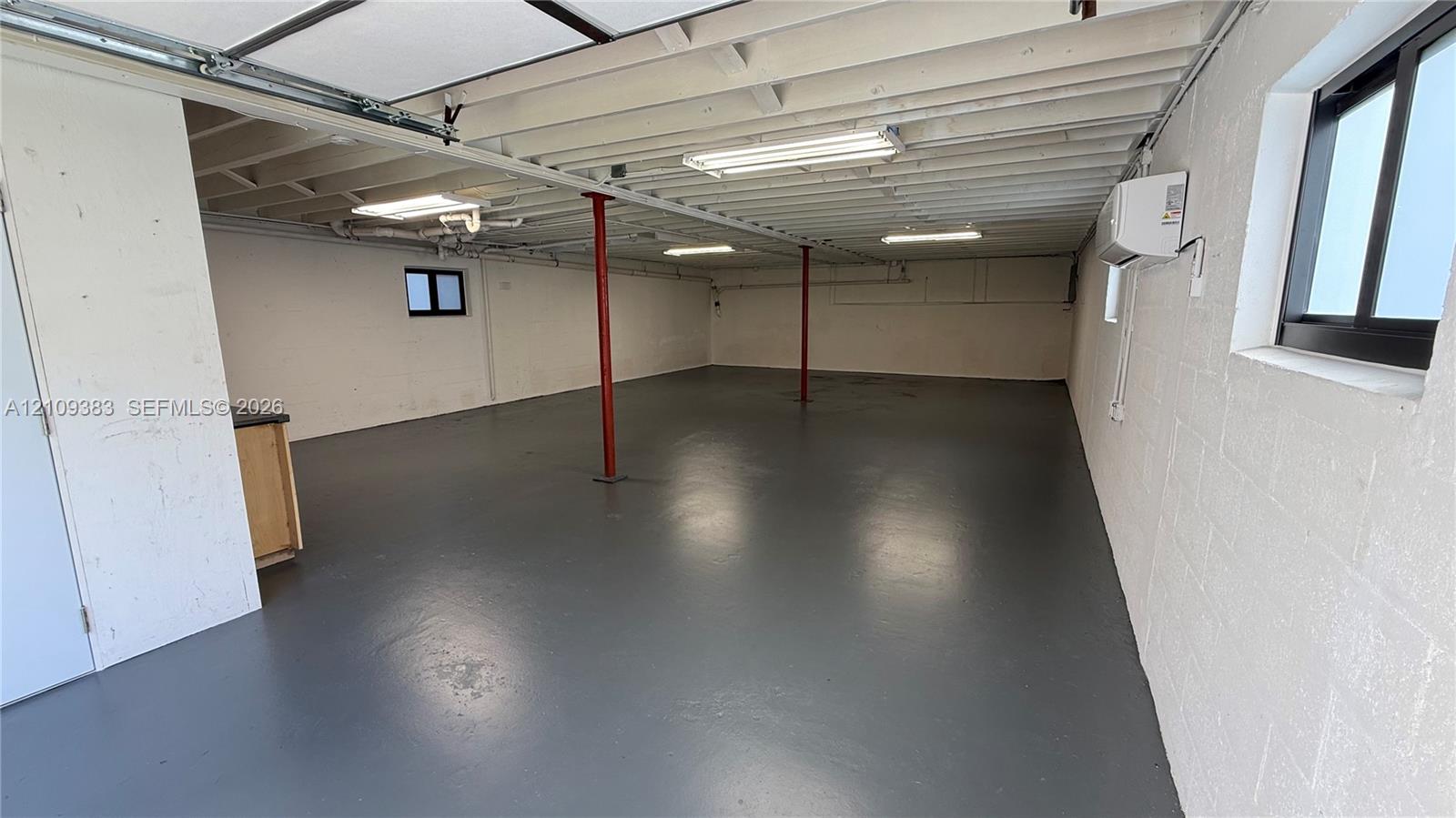 Open interior featuring painted concrete block walls, finished floor surfacing, exposed ceiling joists, overhead fluorescent lighting, and wall-mounted HVAC unit