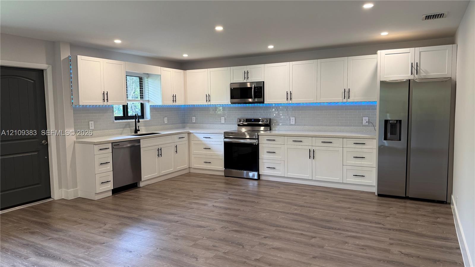Kitchen featuring white shaker-style cabinetry, stainless steel appliances, light-toned stone countertops, subway tile backsplash, and wood-finish flooring