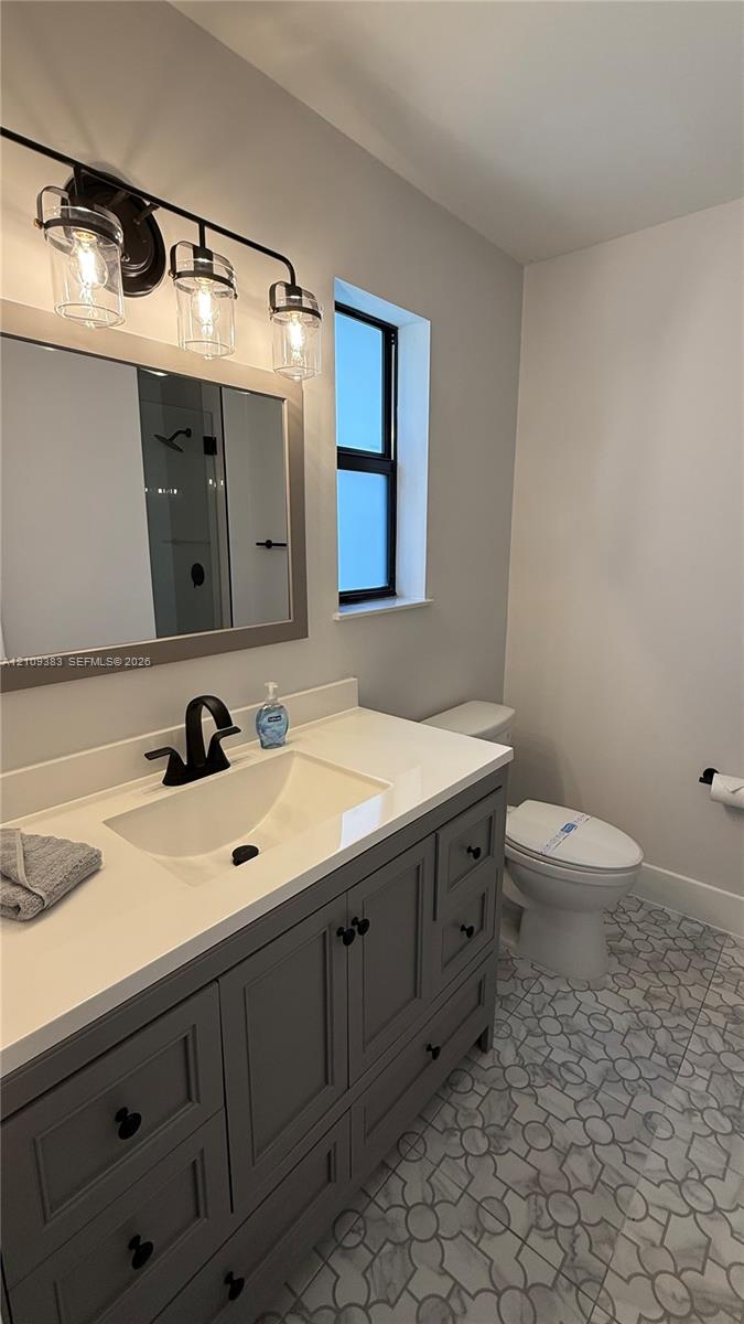 Gray vanity cabinet with a light-toned countertop, undermount sink, matte black faucet, and patterned tile flooring, complemented by a three-light fixture and framed mirror