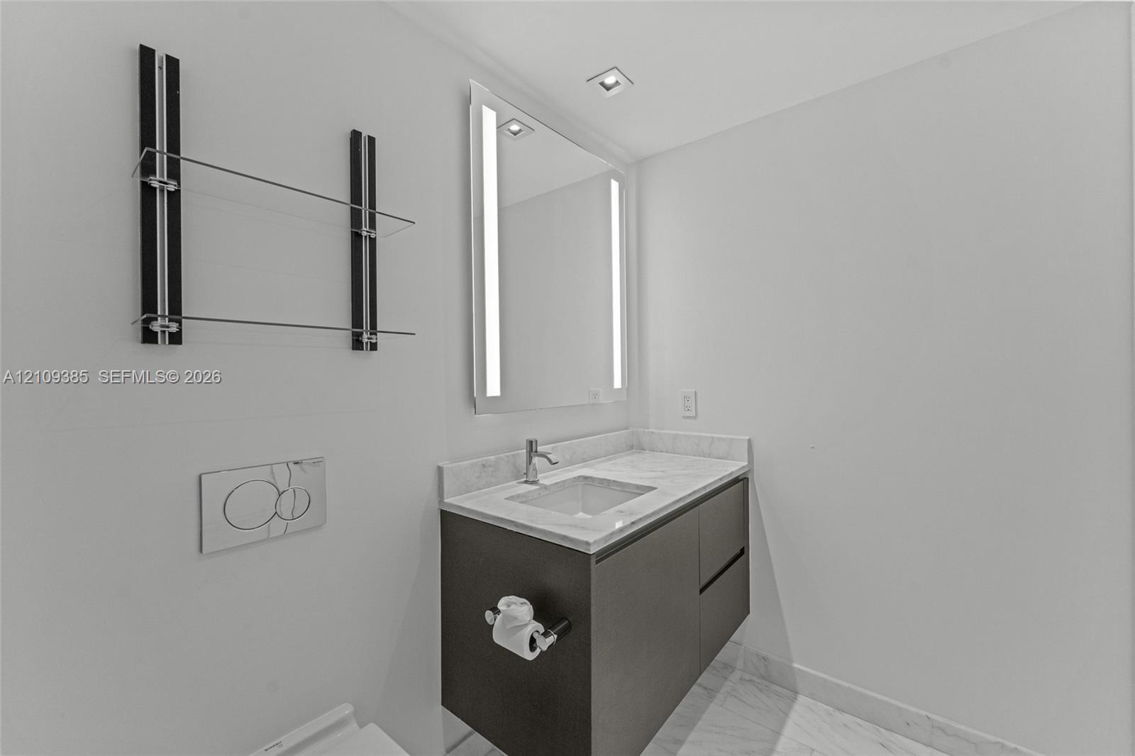 Wall-mounted dark wood-finish vanity with stone countertop, integrated sink, LED-lit mirror, built-in glass shelving, and wall-mounted dual-flush actuator