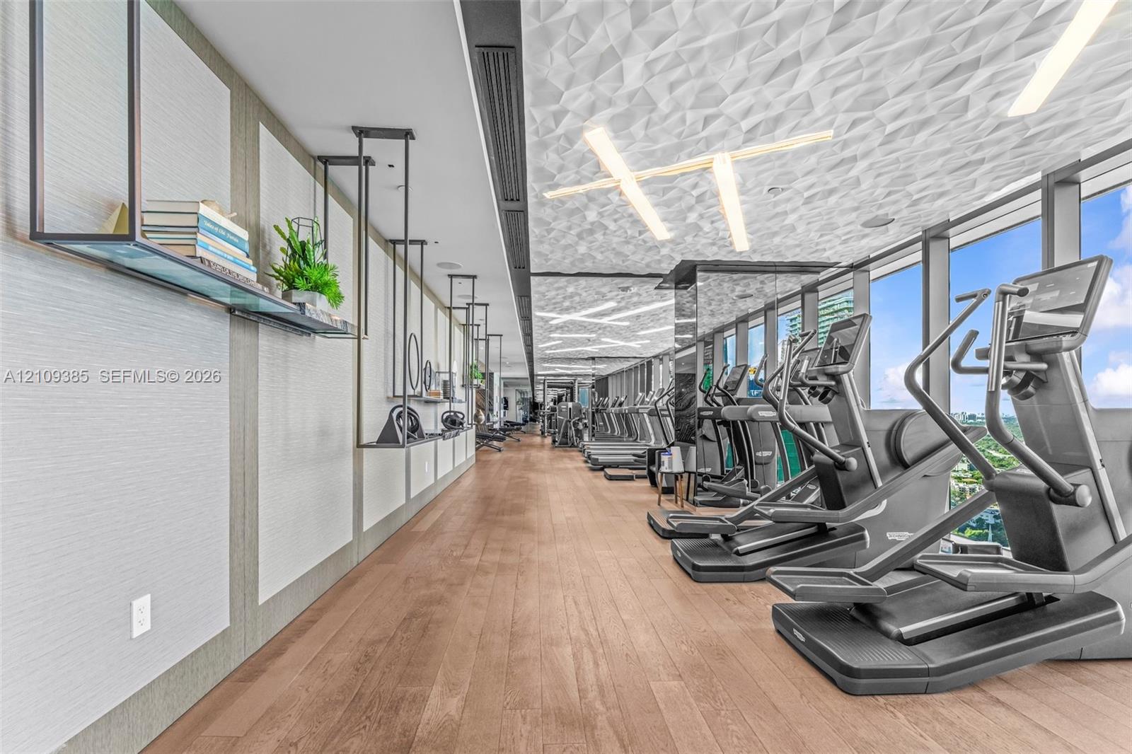 Fitness center featuring wood-finish flooring, sculptural geometric ceiling panels, linear recessed lighting, floor-to-ceiling windows, and wall-mounted metal shelving