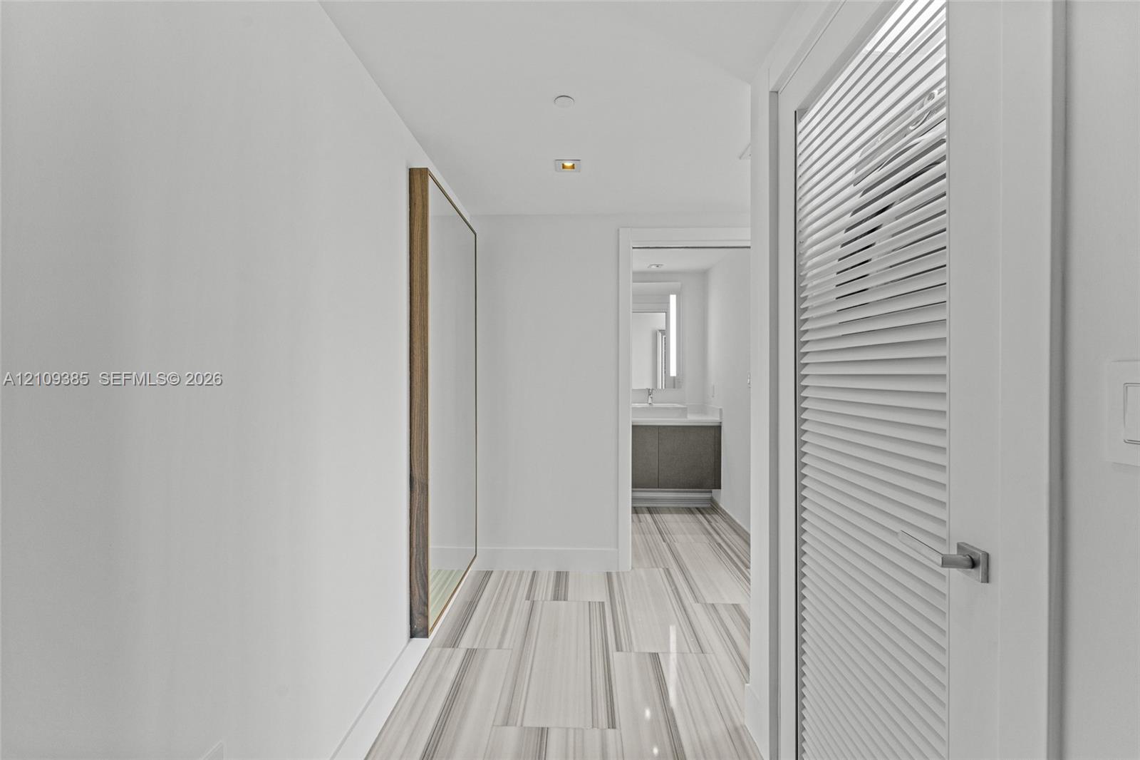 Hallway featuring linear-patterned tile flooring, a louvered white door with satin hardware, recessed ceiling lighting, and a flush-mounted wall mirror with a wood frame