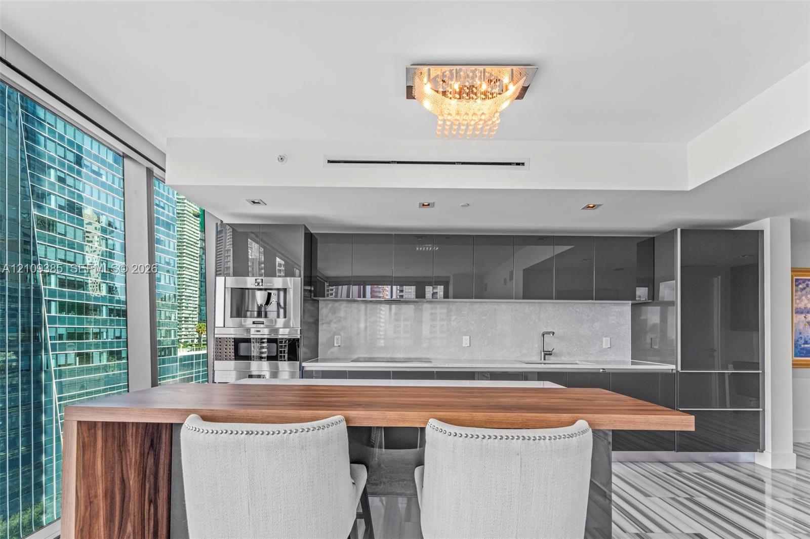 Contemporary kitchen featuring high-gloss cabinetry, stone backsplash, integrated stainless steel coffee system, wood-finish breakfast bar, and polished stone tile flooring