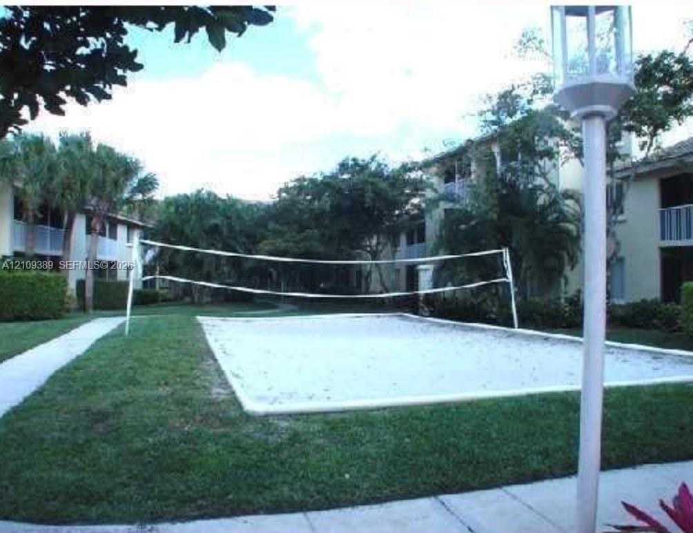 Sand volleyball court featuring a netted playing area, bordered by a manicured lawn, concrete walkways, and mature landscaping