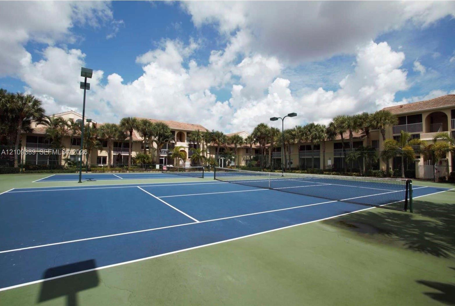 Dual hard-court tennis facility featuring blue and green surfaces, perimeter ball fencing, and integrated pole lighting