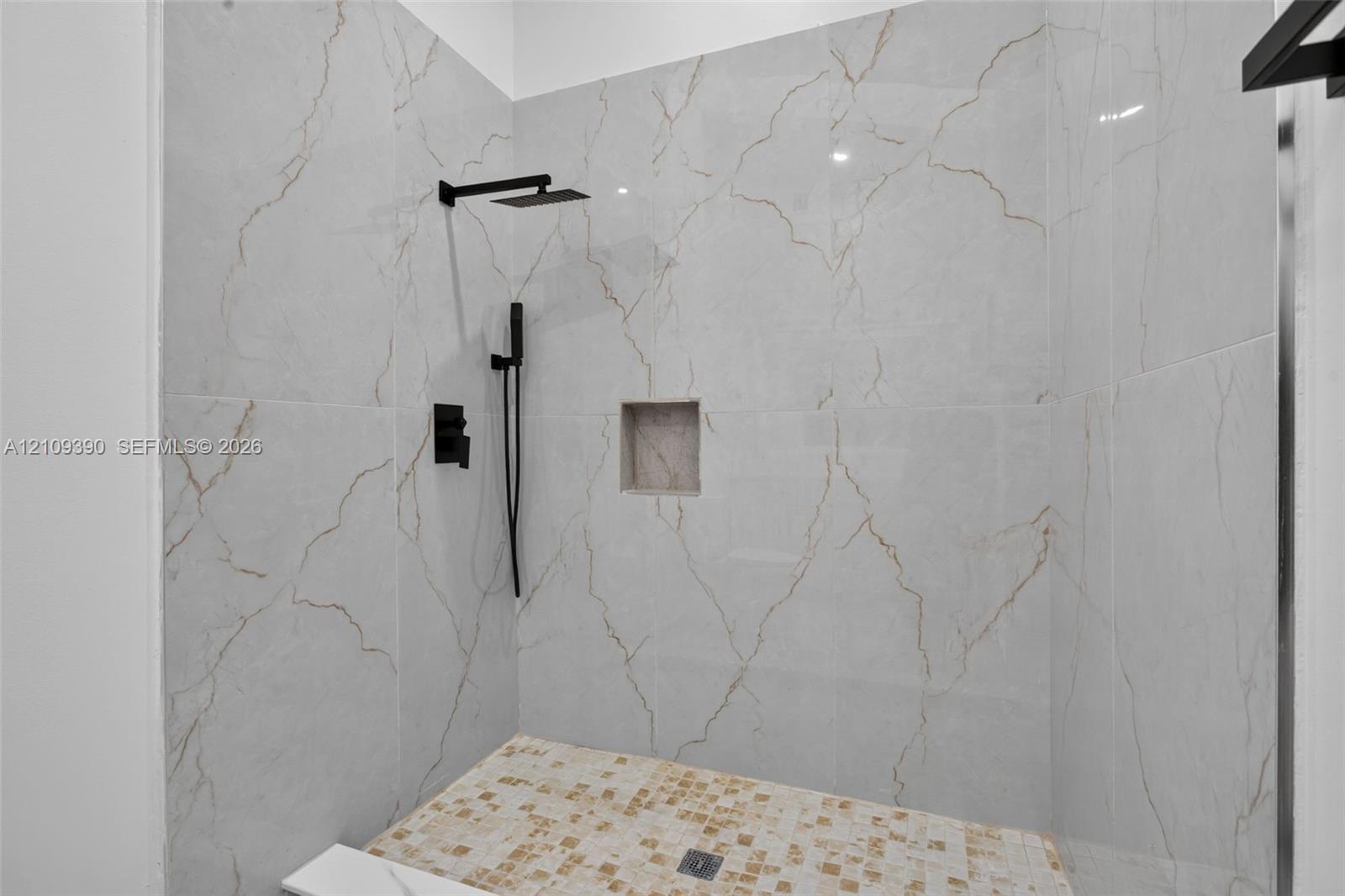 Walk-in shower featuring large-format stone-look wall tiling with veining detail, mosaic tile flooring, built-in recessed wall niche, and matte black rainfall showerhead with hand-held sprayer