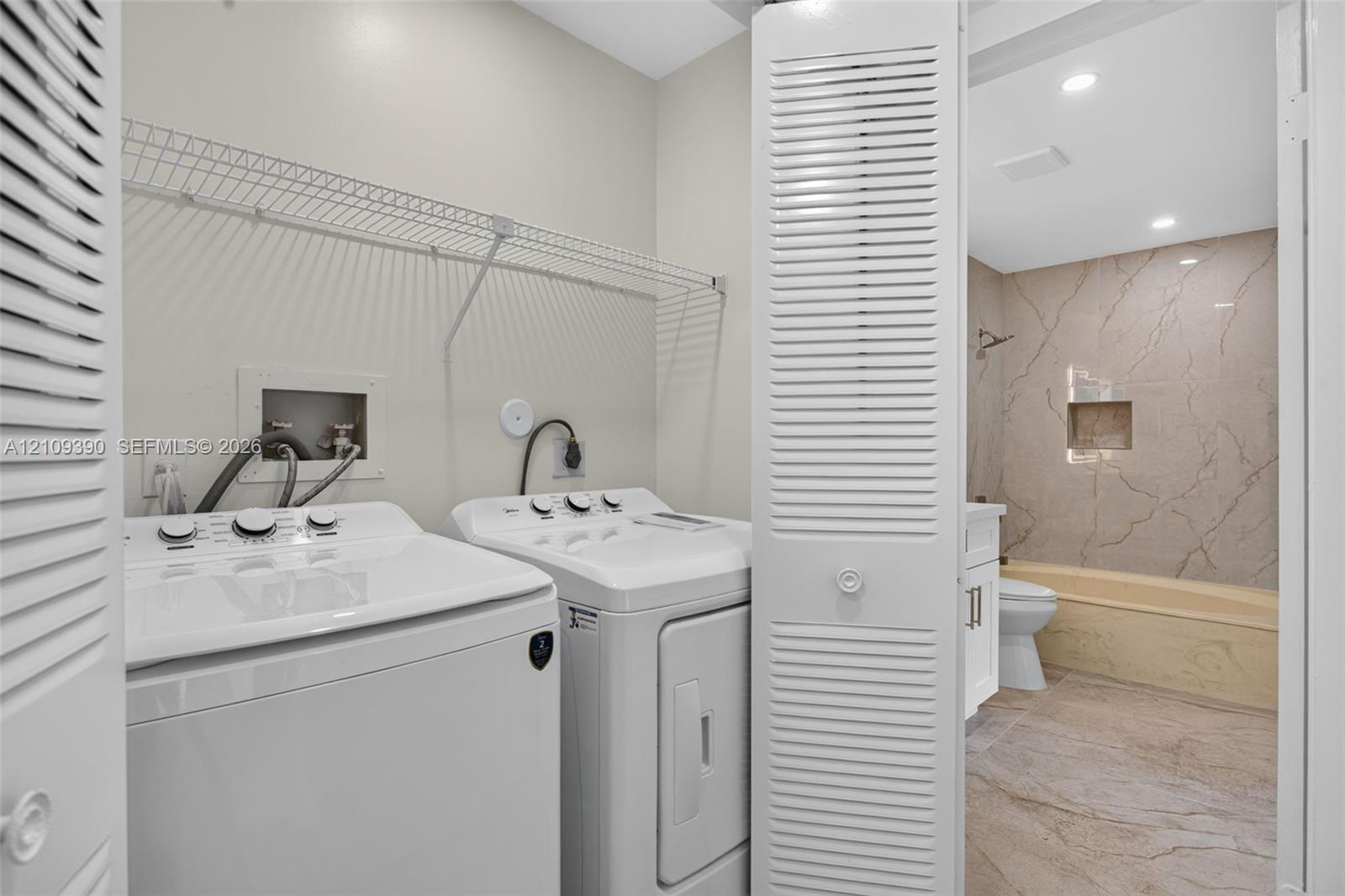 Laundry area featuring wire shelving and accordion-style louvered closet doors, with tile flooring and a tub-shower enclosure finished in a textured stone-look tile