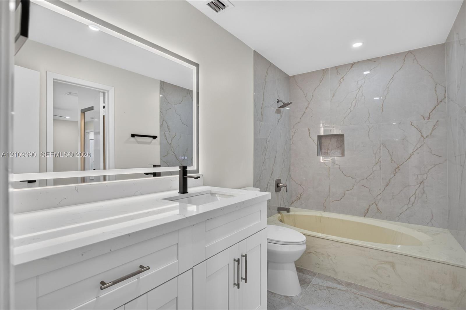 Bathroom featuring a vanity with a white countertop, integrated rectangular sink, matte black faucet, and a large LED-backlit mirror, complemented by a tub enclosure with full-height stone-finish wall tile and a built-in recessed niche