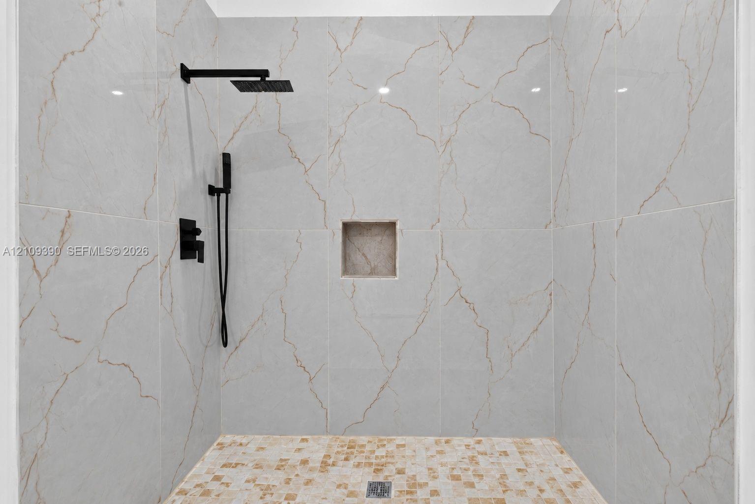 Walk-in shower featuring floor-to-ceiling large-format veined tile, mosaic floor tiling, integrated wall niche, and matte black rainfall showerhead with hand-held sprayer