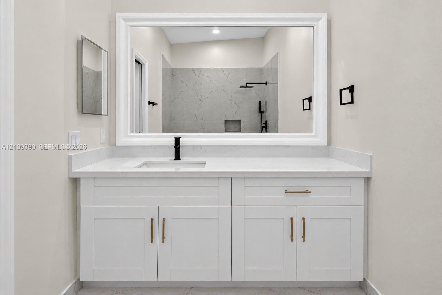 White shaker-style cabinetry with brass hardware, solid surface countertop, rectangular undermount sink, matte black faucet, and large framed wall mirror