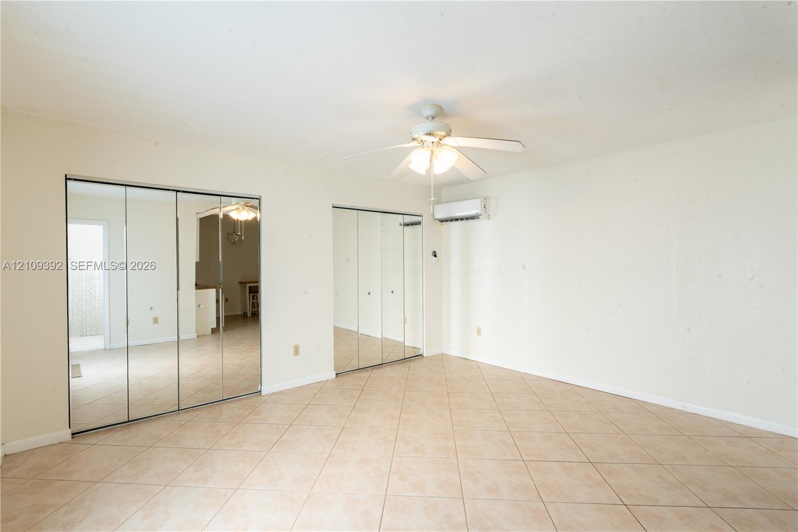 Neutral-toned tile flooring, dual mirrored sliding-door closets, ceiling fan with integrated lighting, and wall-mounted climate control unit