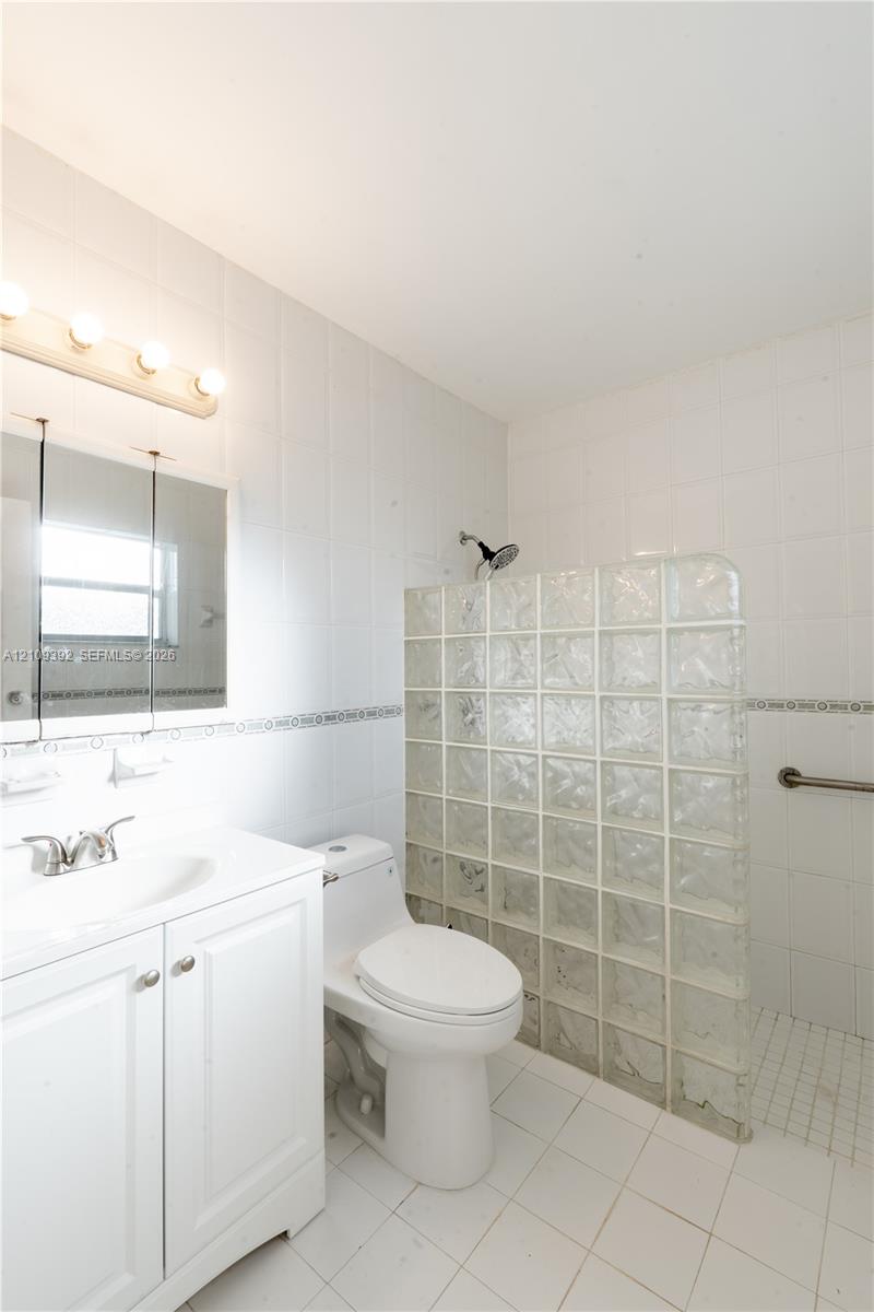 Bathroom featuring a white vanity cabinet, ceramic tile walls and flooring, a glass block shower wall, and a chrome-finish shower head