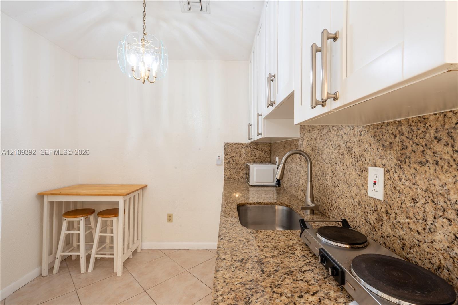 Kitchenette featuring white cabinetry with brushed metal hardware, stone-finish countertops, stainless steel sink with high-arc faucet, integrated electric cooktop, and tile flooring