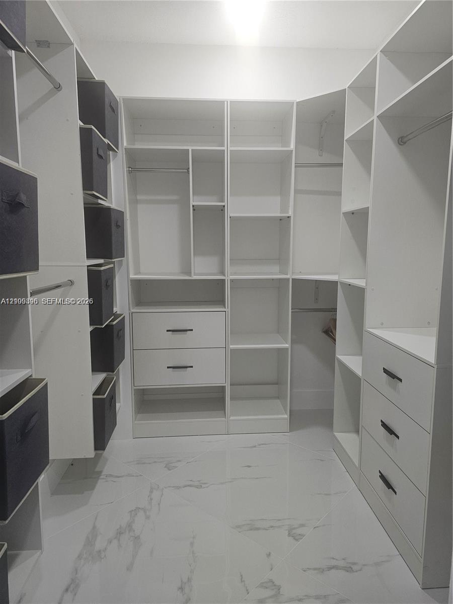 Walk-in closet featuring custom white cabinetry, built-in shelving, integrated dresser drawers with matte black pulls, and large-format polished flooring with a marble-finish aesthetic
