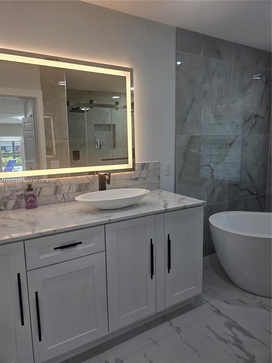 Modern white cabinetry with black hardware, stone-surface countertop, elliptical vessel sink, integrated LED mirror, and soaking tub with large-format tile surround