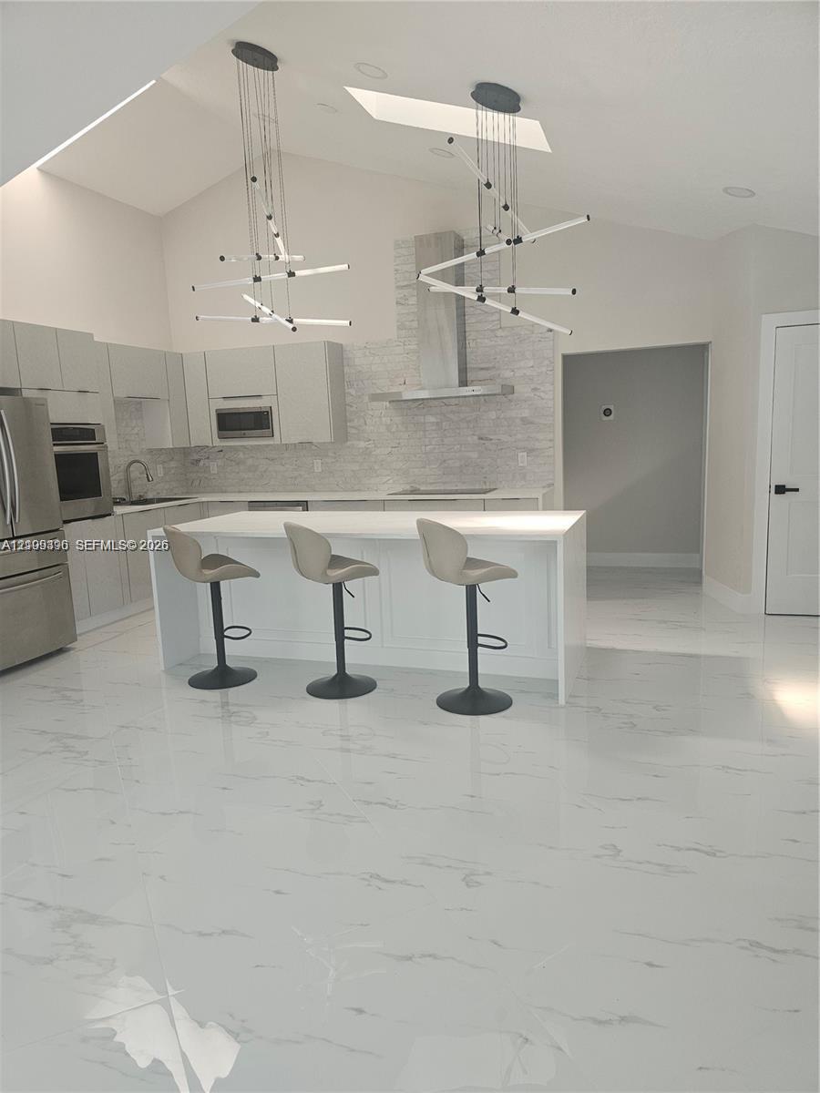 Kitchen featuring vaulted ceilings, skylights, white cabinetry, a center island with solid surfacing, and a stone-tiled backsplash with a stainless steel range hood