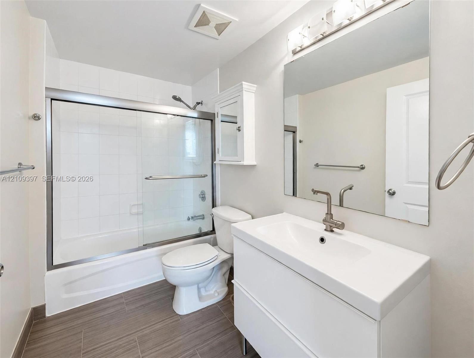 White floating vanity with a ceramic basin, chrome faucet, wall-mounted medicine cabinet, large frameless mirror, and a tub-shower combination featuring a glass sliding enclosure and tile surround