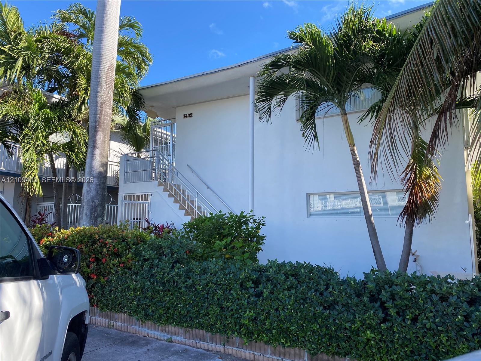 Two-story structure featuring a smooth stucco exterior, integrated metal staircase, horizontal windows, and mature palm landscaping