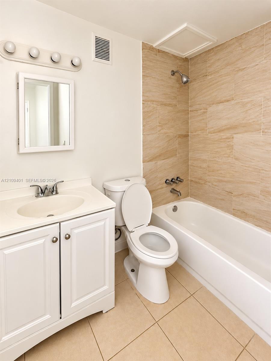White vanity cabinet with integrated basin, polished metal faucet, recessed medicine cabinet, tiled tub surround, and ceramic floor tiling