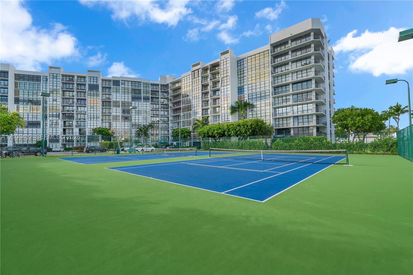 Multi-story residential building exterior featuring twin hard-surface tennis courts with perimeter fencing and integrated lighting