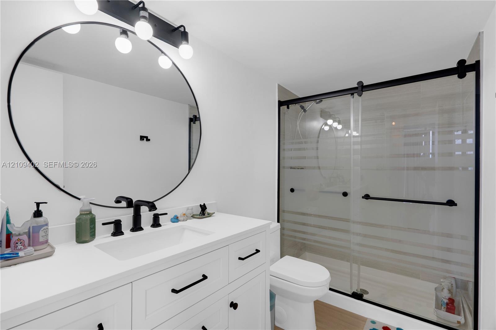 White cabinetry vanity with a solid-surface countertop, matte black hardware, large circular mirror, and walk-in shower featuring a sliding frosted glass door with matte black frame