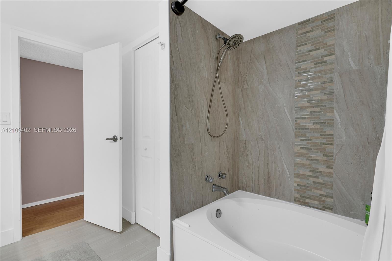 Bathtub featuring a tiled surround with stone-finish panels, vertical mosaic accent inlay, handheld showerhead, and wall-mounted faucet
