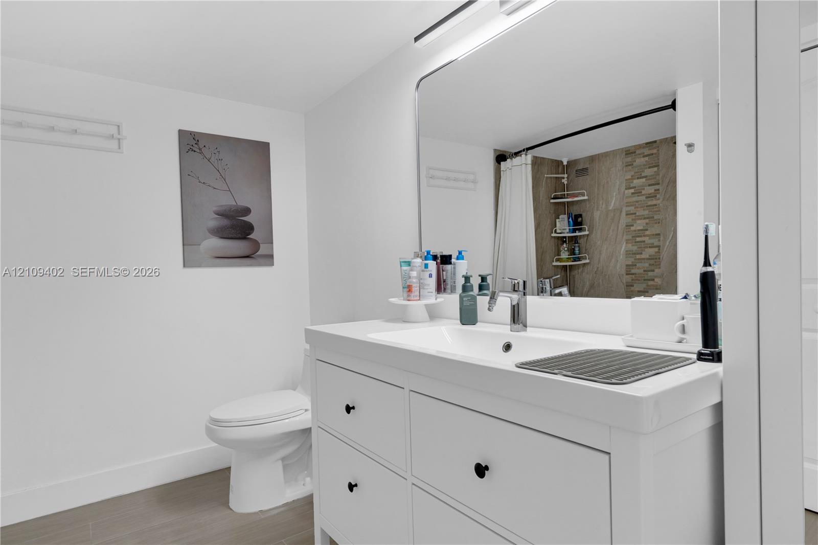 Bathroom featuring a white vanity cabinet with a rectangular basin, large wall-mounted mirror, wood-finish flooring, and a tiled shower surround with decorative stone-finish accents