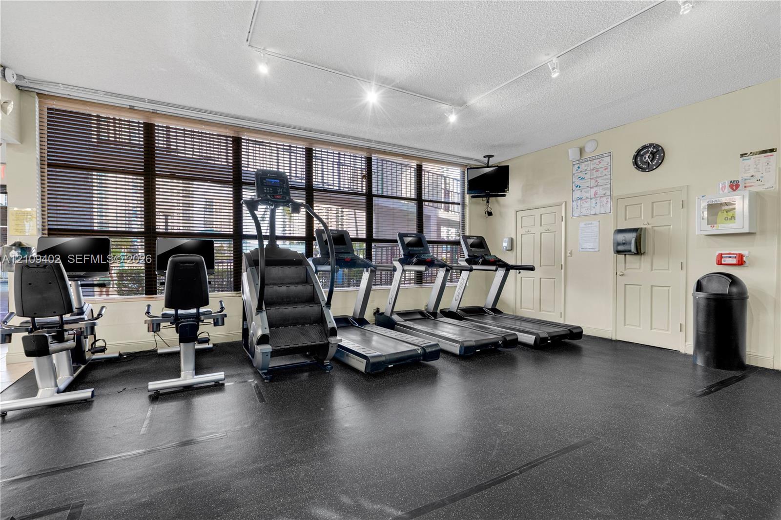 Fitness center featuring wall-to-wall windows with slat blinds, recessed track lighting, and durable rubber flooring