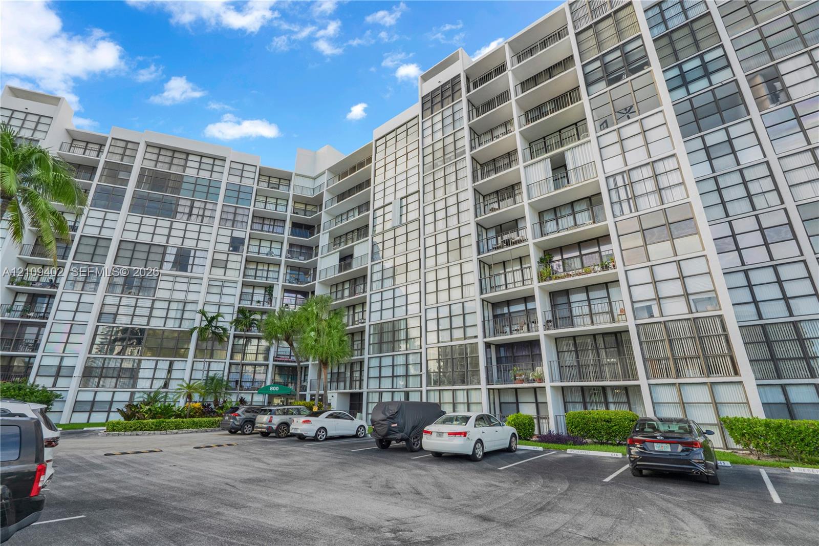 Multi-story residential building featuring a glass-paneled facade, private balconies, and an asphalt surface parking area