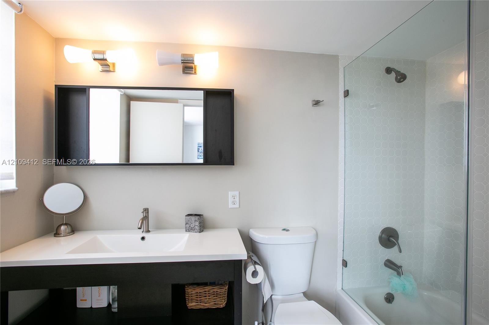 Bathroom featuring a dark-finish vanity with a rectangular sink, white countertop, dual wall-mounted light fixtures, framed medicine cabinet, and a glass-enclosed shower with metallic hardware